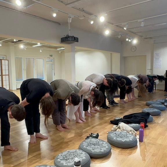 Actors bow in a curtain call line at the conclusion of 'Faith Hope Charity'. Cushions, water bottles, and other accessories are scattered on the floor in front of them. Audience members are pictured to the right of the performers, applauding.